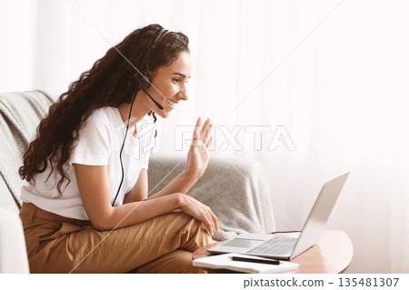 Happy young brunette woman with headset waving to laptop screen, having video call with her family or friends, sitting on sofa at home, side view, copy space. Communication during quarantine Happy young brunette woman with headset waving to laptop screen, having video call with her family or friends, sitting on sofa at home, side view, copy space. Communication during quarantine 135481307