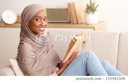 Happy black lady in traditional hijab sitting on comfy sofa with book at home, copy space. Beautiful Muslim lady in Arabic headscarf relaxing on couch, reading interesting story, indoors Happy black lady in traditional hijab sitting on comfy sofa with book at home, copy space. Beautiful Muslim lady in Arabic headscarf relaxing on couch, reading interesting story, indoors 135481309