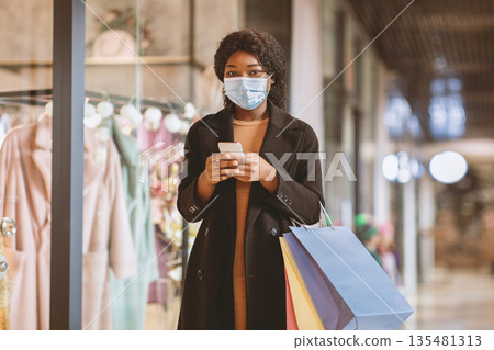 Modern gadgets for shopping and receive messages from store about sales and Black Friday. Young african american woman in protective mask holds shopping bags and uses phone to type on social media Modern gadgets for shopping and receive messages from store about sales and Black Friday. Young african american woman in protective mask holds shopping bags and uses phone to type on social media 135481313