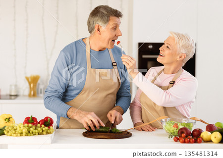 Happy Senior Wife Feeding Husband Cooking Together Standing In Modern Kitchen At Home. Aged Spouses Enjoying Dinner Preparation Tasting Meal Indoors. Elderly Family Nutrition Concept Happy Senior Wife Feeding Husband Cooking Together Standing In Modern Kitchen At Home. Aged Spouses Enjoying Dinner Preparation Tasting Meal Indoors. Elderly Family Nutrition Concept 135481314