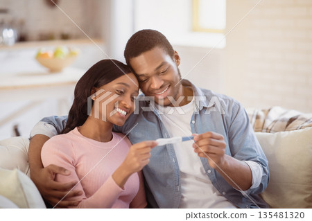 Smiling future mom and dad with positive pregnancy test waiting for their baby at home. Young black man hugging his happy expectant wife indoors, learning happy news together 135481320