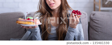 Depressed young lady having eating disorder, comforting herself with sweets on sofa at home. Unhappy millennial woman coping with negative emotions through food, suffering from bulimia Depressed young lady having eating disorder, comforting herself with sweets on sofa at home. Unhappy millennial woman coping with negative emotions through food, suffering from bulimia 135481330