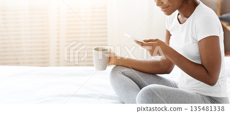 Smiling african american young lady in pajamas drinking morning tea or coffee, using mobile phone, checking social media or reading messages, sitting on bed at home, side view, copy space Smiling african american young lady in pajamas drinking morning tea or coffee, using mobile phone, checking social media or reading messages, sitting on bed at home, side view, copy space 135481338