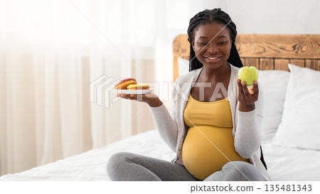 Pregnancy Diet. Smiling Pregnant Woman Choosing Green Apple Instead Of Donuts, Enjoying Organic Vitamin Fruits And Healthy Nutrition, Joyful African American Expectant Mother Sitting On Bed At Home Pregnancy Diet. Smiling Pregnant Woman Choosing Green Apple Instead Of Donuts, Enjoying Organic Vitamin Fruits And Healthy Nutrition, Joyful African American Expectant Mother Sitting On Bed At Home 135481343