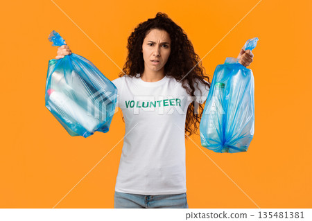Displeased Female Volunteer Holding Two Plastic Garbage Bags Full Of Litter And Wasted Bottles Posing On Yellow Studio Background. Junk Sorting, Ecology Concept 135481381