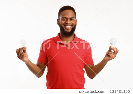 African American Guy Holding Two Light Bulbs Standing On White Studio Background, Choosing Energy-Saving Lightbulb. Save Energy, Safe Light-Bulbs Disposal Concept 135481390