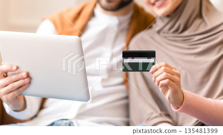 Muslim Couple Shopping Online With Credit Card And Digital Tablet Computer Sitting On Couch At Home, Focus On Bank Card. Happy Customers, Internet Shop Sales And Discounts Concept. Shallow Depth 135481399