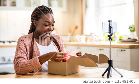 Unboxing Vlog. Black Millennial Influencer Woman Filming Video Content For Her Blog, Smiling African Female Blogger Unpacking New Clothes In Front Of Camera, Using Smartphone On Tripod, Free Space Unboxing Vlog. Black Millennial Influencer Woman Filming Video Content For Her Blog, Smiling African Female Blogger Unpacking New Clothes In Front Of Camera, Using Smartphone On Tripod, Free Space 135481405