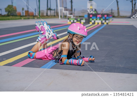 A girl in a bright pink helmet, protective elbow pads and roller skates lies on the skatepark path A girl in a bright pink helmet, protective elbow pads and roller skates lies on the skatepark path 135481421