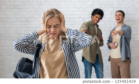 Bullying and peer relationships. Focus on blonde teen lady with backpack covering her ears, not to hear taunts from multiracial male students in school or college, on white brick wall background 135481429