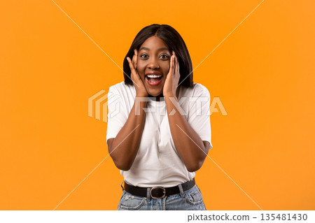 Emotional black lady in white t-shirt screaming and touching her face over orange studio background, emotional african american young woman expressing amazement, seeing something exciting 135481430