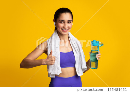 Stay Hydrated. Portrait of happy smiling female fitness instructor in wireless earphones holding bottle with fresh water and showing thumbs up sign gesture, posing isolated on orange studio background 135481431