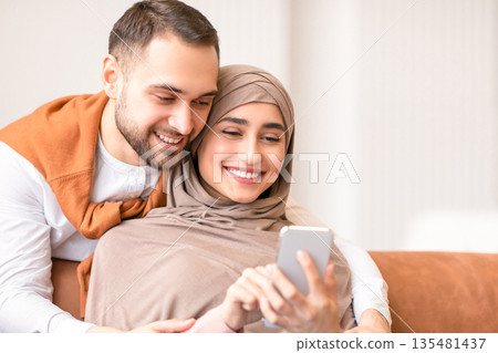 Happy Muslim Couple Using Smartphone Texting And Browsing Internet Sitting On Couch At Home. Family Leisure. Cheerful Arab Spouses Watching Video Online On Phone. Great Mobile Application Concept 135481437
