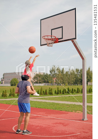 A girl sits on her father's shoulders, throwing a basketball into the ring. Family basketball game in the outdoor stadium on a sunny summer day A girl sits on her father's shoulders, throwing a basketball into the ring. Family basketball game in the outdoor stadium on a sunny summer day 135481531
