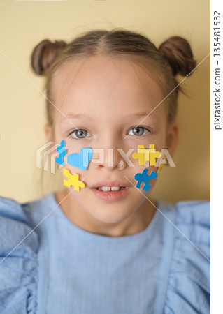 Portrait of a girl with clear eyes, ornaments on her cheek in the shape of hearts and pieces of yellow and blue puzzles. Awareness and social support for children with down syndrome 135481532