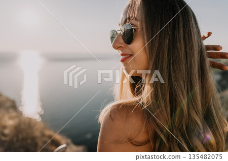 Woman sunset coastline smiling young woman wearing sunglasses looks at the ocean during golden hour while enjoying a summer vacation adventure Woman sunset coastline smiling young woman wearing sunglasses looks at the ocean during golden hour while enjoying a summer vacation adventure 135482075