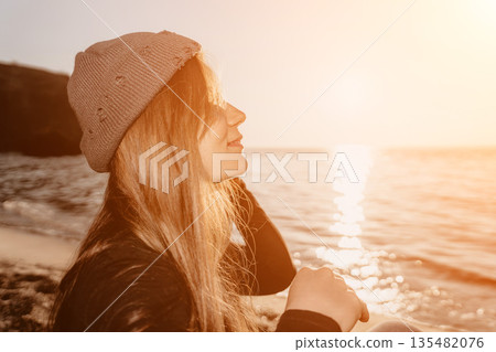 Woman, sunset, beach. Young blonde girl wearing a beanie hat sitting by the ocean, enjoying the serene golden hour light. Woman, sunset, beach. Young blonde girl wearing a beanie hat sitting by the ocean, enjoying the serene golden hour light. 135482076