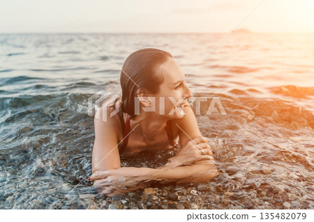 Woman, beach, ocean: happy woman relaxes in shallow sea water enjoying golden hour sunset Woman, beach, ocean: happy woman relaxes in shallow sea water enjoying golden hour sunset 135482079