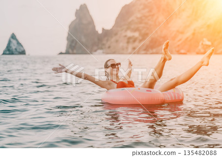 Woman, floating, donut. Joyful woman floats on inflatable donut in open sea with rocky cliffs, enjoying summer leisure. 135482088