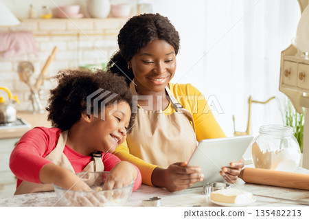 Happy black mom and her little daughter checking dough recipe online on digital tablet while baking pastry in kitchen together, young family enjoying modern technologies and homemade food, free space Happy black mom and her little daughter checking dough recipe online on digital tablet while baking pastry in kitchen together, young family enjoying modern technologies and homemade food, free space 135482213