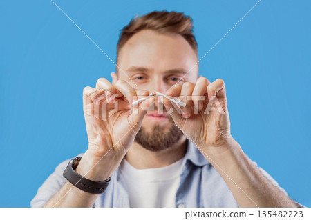 Stop smoking concept. Young bearded guy breaking cigarette on blue studio background, selective focus. Millennial man fighting his addiction to nicotine, saying NO to harmful habit 135482223