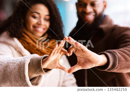 Celebrating Valentine's Day. Close up of smiling loving black man and woman making heart shape with fingers walking together in the evening, looking at hands, selective focus, blurred background Celebrating Valentine's Day. Close up of smiling loving black man and woman making heart shape with fingers walking together in the evening, looking at hands, selective focus, blurred background 135482250
