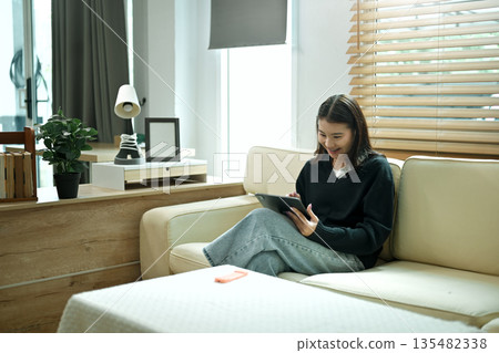A young woman relaxing on a sofa using a tablet at home, enjoying a calm holiday mood with comfort and leisure time A young woman relaxing on a sofa using a tablet at home, enjoying a calm holiday mood with comfort and leisure time 135482338
