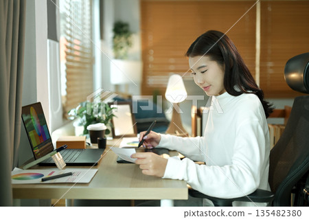 Female graphic designer working on logo creation with drawing tablet and color palette in modern bright office. 135482380
