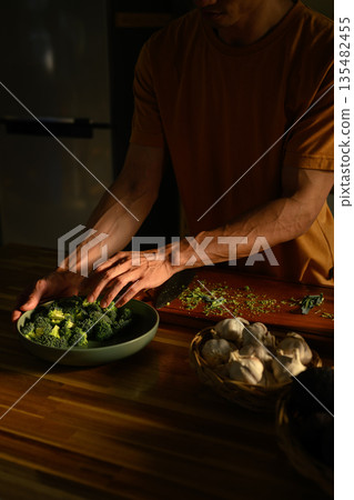 Man preparing vegetables in a kitchen, placing broccoli into a bowl, healthy eating, care, and nutrition after daily activity 135482455