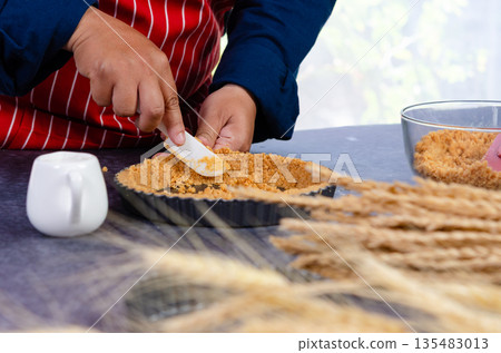Baker wearing red and white striped apron spreading golden graham cracker crumbs into fluted tart pan using white spatula on kitchen countertop Baker wearing red and white striped apron spreading golden graham cracker crumbs into fluted tart pan using white spatula on kitchen countertop 135483013