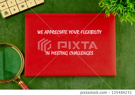 A red card with text We appreciate your flexibility in meeting challenges rests on a green surface with a calculator, magnifying glass, and plant A red card with text We appreciate your flexibility in meeting challenges rests on a green surface with a calculator, magnifying glass, and plant 135484231