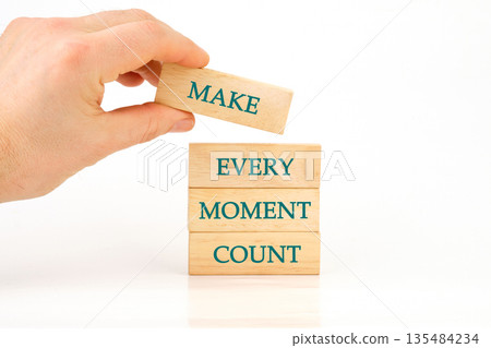 A hand places a wooden block with the word Make on top of a stack. The stack of blocks reads Every Moment Count against a clean, white backdrop 135484234