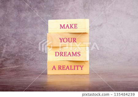 stacked wooden blocks with pink lettering spell out make your dreams a reality. the blocks are sitting on a wood surface in front of a grey background stacked wooden blocks with pink lettering spell out make your dreams a reality. the blocks are sitting on a wood surface in front of a grey background 135484239