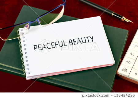 A spiral notebook with the words Peaceful Beauty is on a closed, green book. Eyeglasses, a pen, and a calculator are nearby on a dark red surface A spiral notebook with the words Peaceful Beauty is on a closed, green book. Eyeglasses, a pen, and a calculator are nearby on a dark red surface 135484256