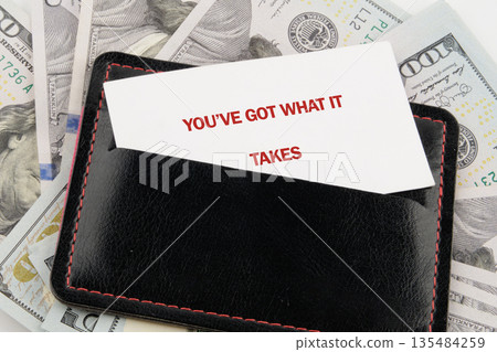 A black leather wallet holds a white business card that reads YOUVE GOT WHAT IT TAKES. The wallet is placed on top of several United States one hundred dollar bills 135484259