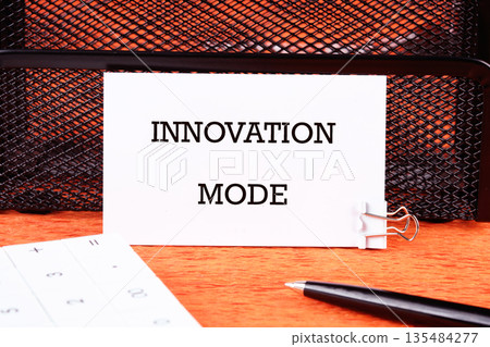White paper says Innovation Mode. A pen and calculator sit on a wood surface. A black mesh container is behind the paper 135484277