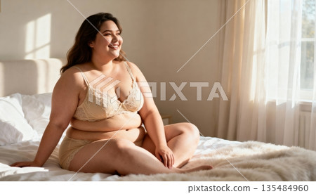A happy young adult plus-size woman wearing beige lace lingerie sits comfortably on a white bed, smiling and looking away in a sunlit bedroom. 135484960