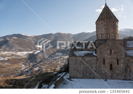 Ancient stone architecture of Tatev Monastery stands against snowy mountain slopes under clear winter light, warm stone textures 135485642