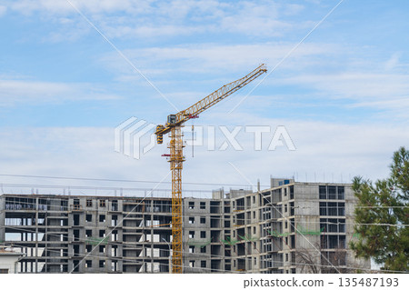 Construction process of multi-storey building from monolithic frame of reinforced concrete panels using orange tower-type construction crane against background of partially cloudy blue sky in city Construction process of multi-storey building from monolithic frame of reinforced concrete panels using orange tower-type construction crane against background of partially cloudy blue sky in city 135487193