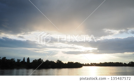 Witness the breathtaking spectacle of clouds as they dance across the sky, casting shadows over calm waters at sunset. Natures palette transitions with stunning hues of orange and blue 135487572