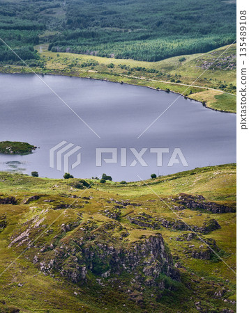 A lake stretches out surrounded by green hills and trees. Rocks form a terrace in the foreground. The day appears cloudy. A lake stretches out surrounded by green hills and trees. Rocks form a terrace in the foreground. The day appears cloudy. 135489108