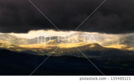 Sunlight filters through a gap in dark, stormy clouds, illuminating sections of mountainous terrain in National Park, creating a dramatic vista. Sunlight filters through a gap in dark, stormy clouds, illuminating sections of mountainous terrain in National Park, creating a dramatic vista. 135489121