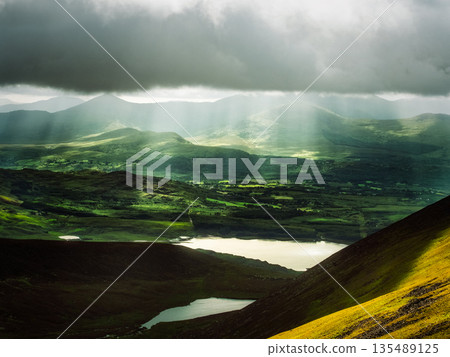 Sunlight streams through dark clouds, illuminating the green hills and fields of Killarney National Park. Lakes reflect the light in the foreground. Sunlight streams through dark clouds, illuminating the green hills and fields of Killarney National Park. Lakes reflect the light in the foreground. 135489125