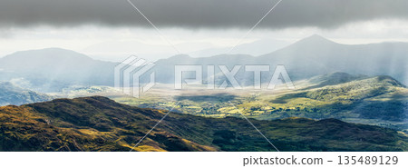 Mountainous landscape shows a valley in County Kerry, Ireland. A cloud covered sky filters sunlight, creating rays over the green land. Mountains rise in the background. 135489129
