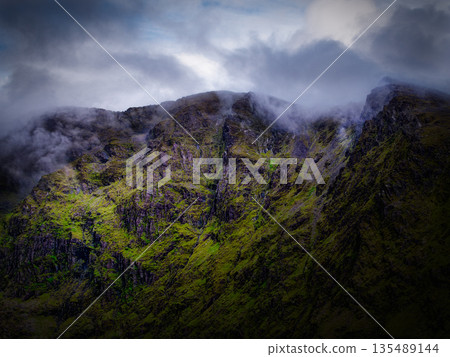 Dramatic landscape with steep mountain side partly hidden by low clouds. Green vegetation clings to the rocks under soft, natural light. 135489144