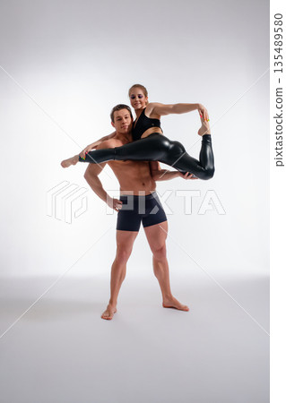 Acrobatic duo performs a graceful balancing pose. A strong man supports a flexible woman against a white studio background. 135489580