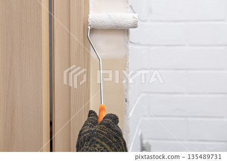 A woman wearing a glove paints a wall with white paint using a roller 135489731