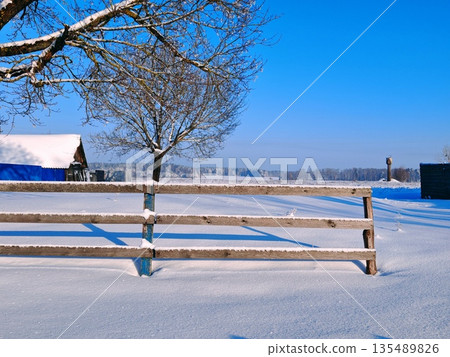 Snow Covered Wooden Fence Rural Winter. Rustic wooden fence covered in fresh snow with bare tree and distant snowy fields under bright blue sky. Cozy winter countryside scene and rural charm. Snow Covered Wooden Fence Rural Winter. Rustic wooden fence covered in fresh snow with bare tree and distant snowy fields under bright blue sky. Cozy winter countryside scene and rural charm. 135489826