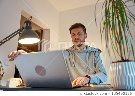 Man standing at home office desk using laptop under desk lamp in apartment interior Man standing at home office desk using laptop under desk lamp in apartment interior 135490136