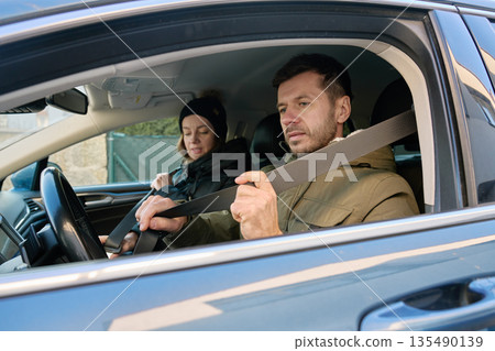 Man fastening seat belt in driver seat while woman sits in passenger seat inside car Man fastening seat belt in driver seat while woman sits in passenger seat inside car 135490139